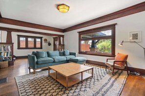 Ein Wohnzimmer mit einer Eckcouch, einem Holztisch, einem gemusterten Teppich, einem Bücherregal und einem Fenster mit Blick ins Grüne.
