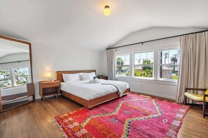 Ein Schlafzimmer mit einem hölzernen Bett, einem roten Teppich mit Muster, einem Fenster mit Vorhängen und einem Sessel.