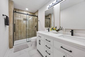 Un baño moderno con cabina de ducha de vidrio, un tocador blanco con pomos negros y un espejo grande.