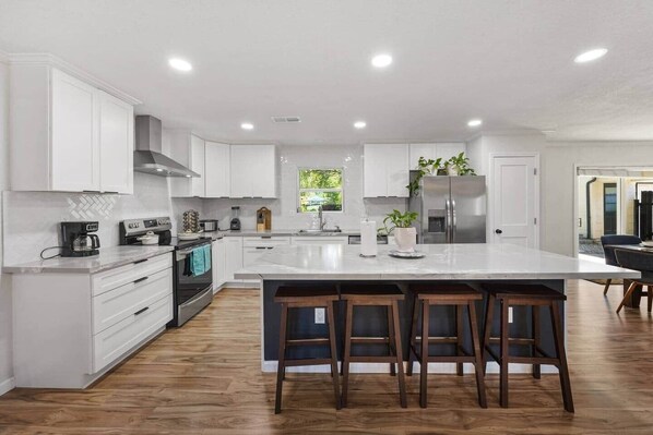 Una cocina moderna con armarios blancos, una isla central con taburetes y un horno empotrado, además de un refrigerador de acero inoxidable.