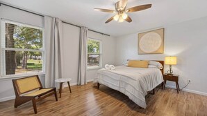 Una camera da letto con un letto, una sedia, un comodino con lampada, una ventola a soffitto e ampie finestre con tende.