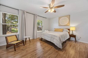 A bedroom with a bed, a chair, a bedside table with a lamp, a ceiling fan, and large windows with curtains.