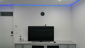 A modern bathroom with a large flat-screen TV on a white cabinet, a clock on the wall, and blue LED lighting.