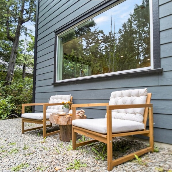 Un espace de détente extérieur moderne, avec deux fauteuils confortables et une table en bois, situé devant une maison dotée d’une grande fenêtre.