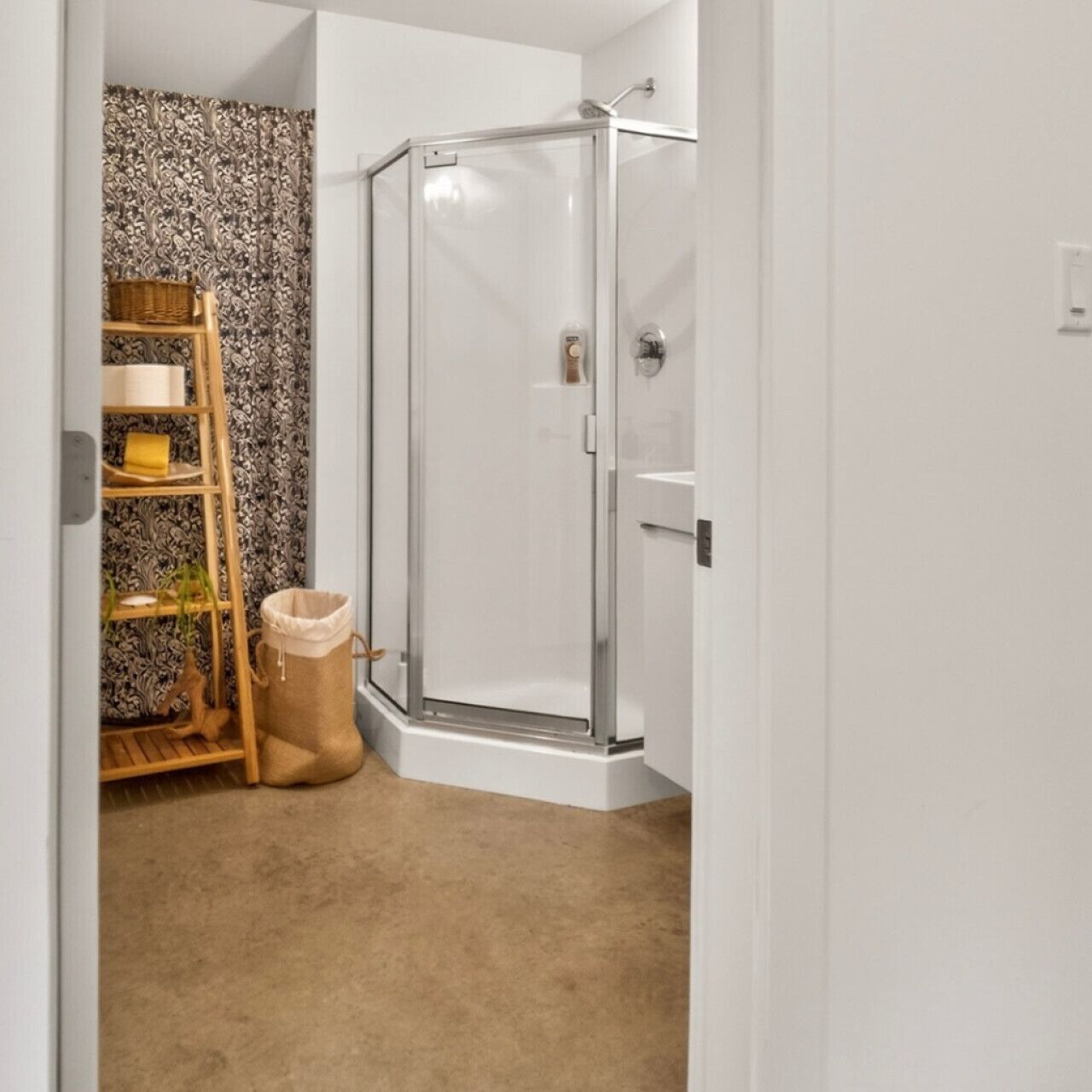 A corner bathroom with a glass shower enclosure, a wooden shelf with baskets, and a beige carpet.
