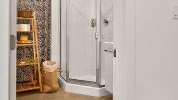 A corner bathroom with a glass shower enclosure, a wooden shelf with baskets, and a beige carpet.