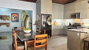 A modern kitchen with a central island, a dining table with chairs, and surfboards on the wall. - 4 Bedroom Oceanview Haven (Dominical)