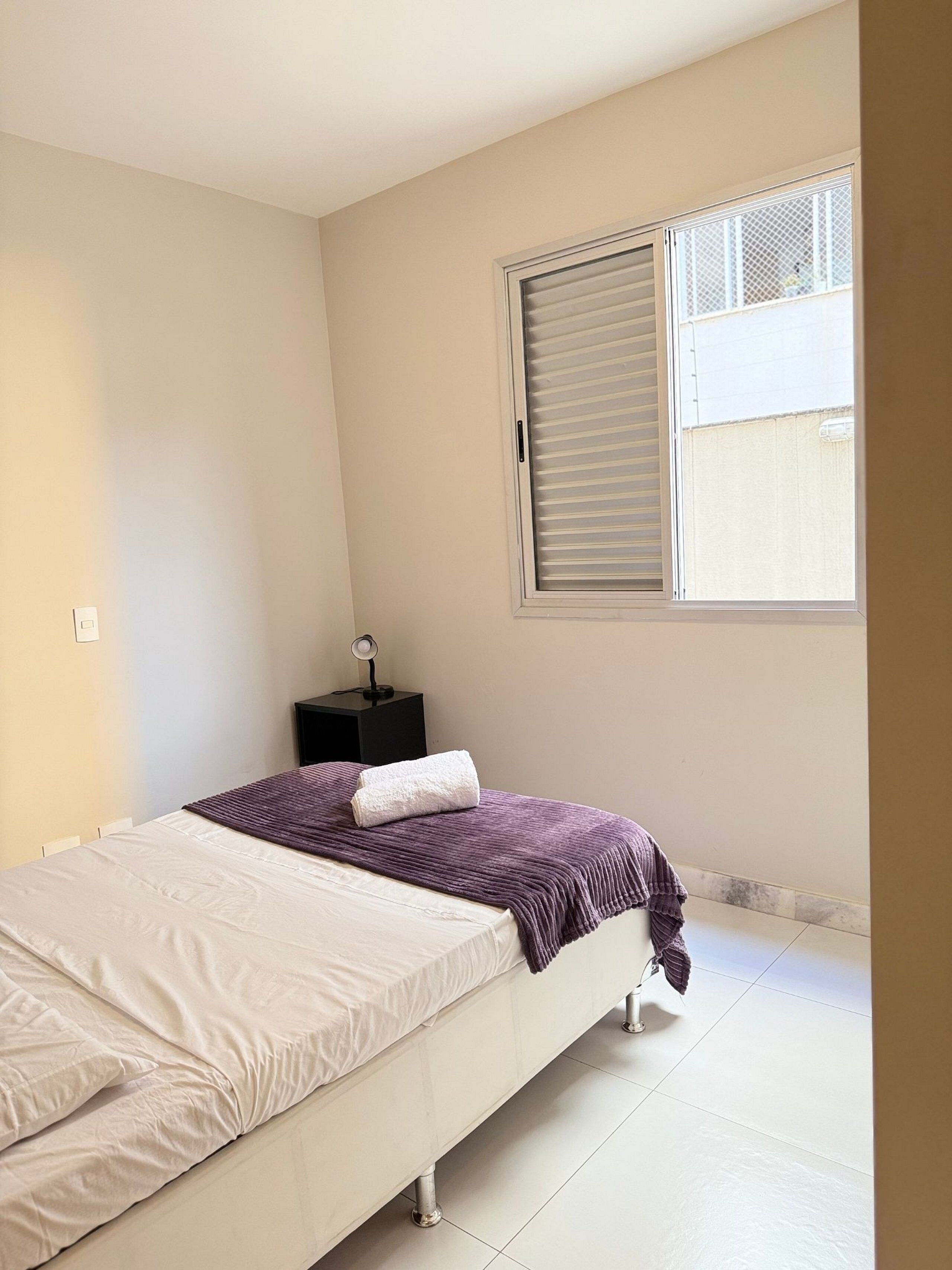Un dormitorio con cama, mesita de noche, ventana con persianas y vistas al exterior.