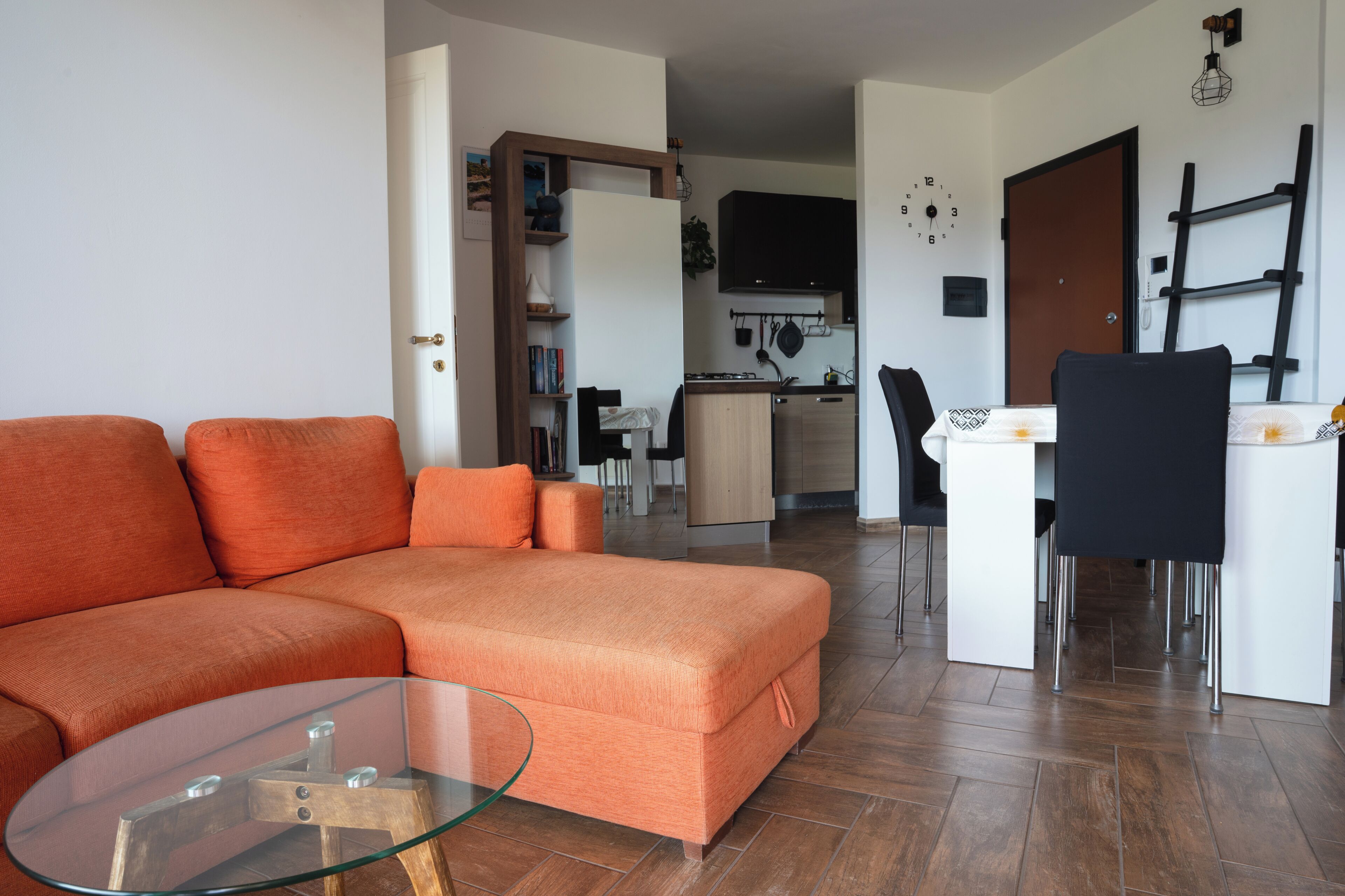Un salon comprenant un canapé d’angle, une table basse en verre, un coin cuisine avec des placards et une cuisinière, et un coin repas avec une table et des chaises.