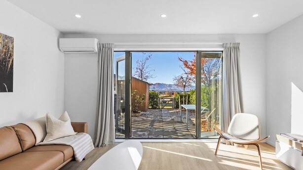Een moderne kamer met een leren bank, een witte stoel, een groot raam met uitzicht op een tuin en bergen, en een houten vloer.