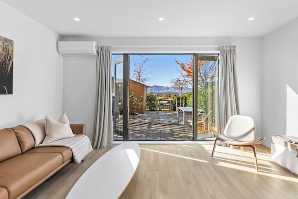 Ein modernes Zimmer mit einer Ledersofa, einem weißen Sessel, einem großen Fenster mit Blick auf einen Garten und Berge sowie einem Holzboden.