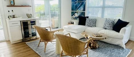A modern living room with a white sofa, wicker chairs, a coffee table, and a large window with blinds.
