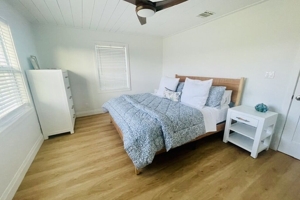 Une chambre avec un plancher en bois, un lit recouvert d’une couvre-lit à motifs, une table de chevet blanche et une commode blanche.