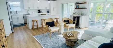 A modern kitchen with white cabinets, a dining area with wicker chairs, and a large window overlooking a deck.