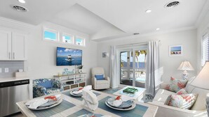 A well-lit living area with a dining table set for two, a sofa, a TV, and a view of the sea through a glass door.