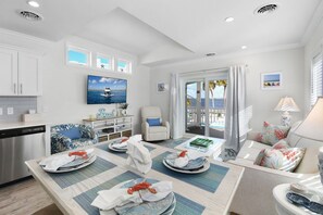 A well-lit living area with a dining table set for two, a sofa, a TV, and a view of the sea through a glass door.