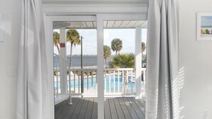 A room with a view of a pool and palm trees, a white curtain, and a white wall with a picture.