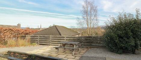 Un terraç de fusta amb taula de picnic, un jardí elevat i vista a unes colines i una església al fons.