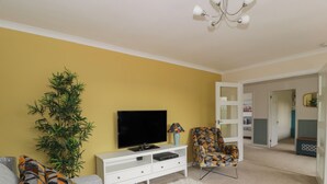 Una sala de estar con un sofá, un televisor, un sillón estampado, un armario blanco, una alfombra mullida y una puerta que da a otra habitación.