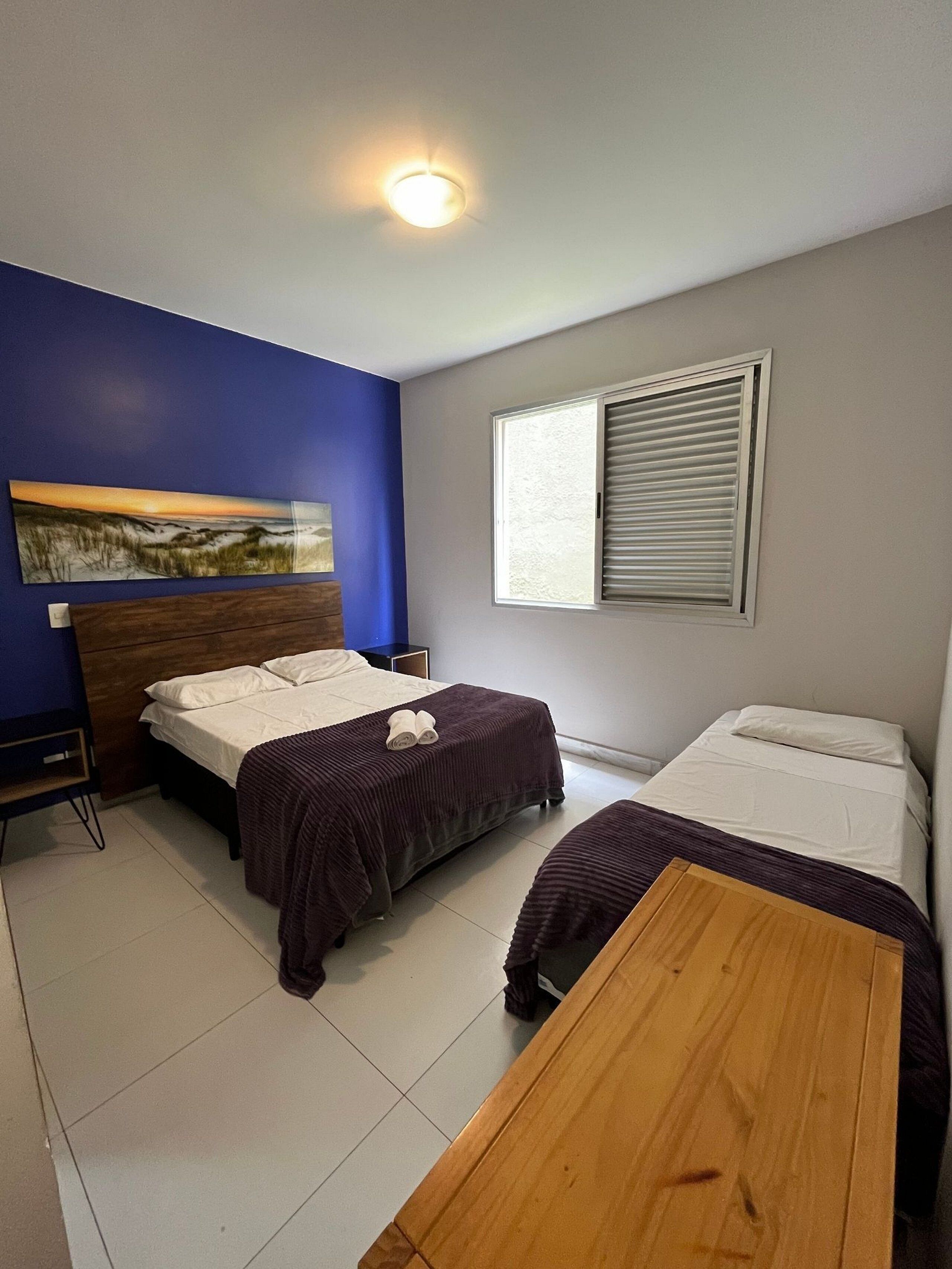 A room with two beds, a wooden table, a painting on the wall, and a window with blinds.
