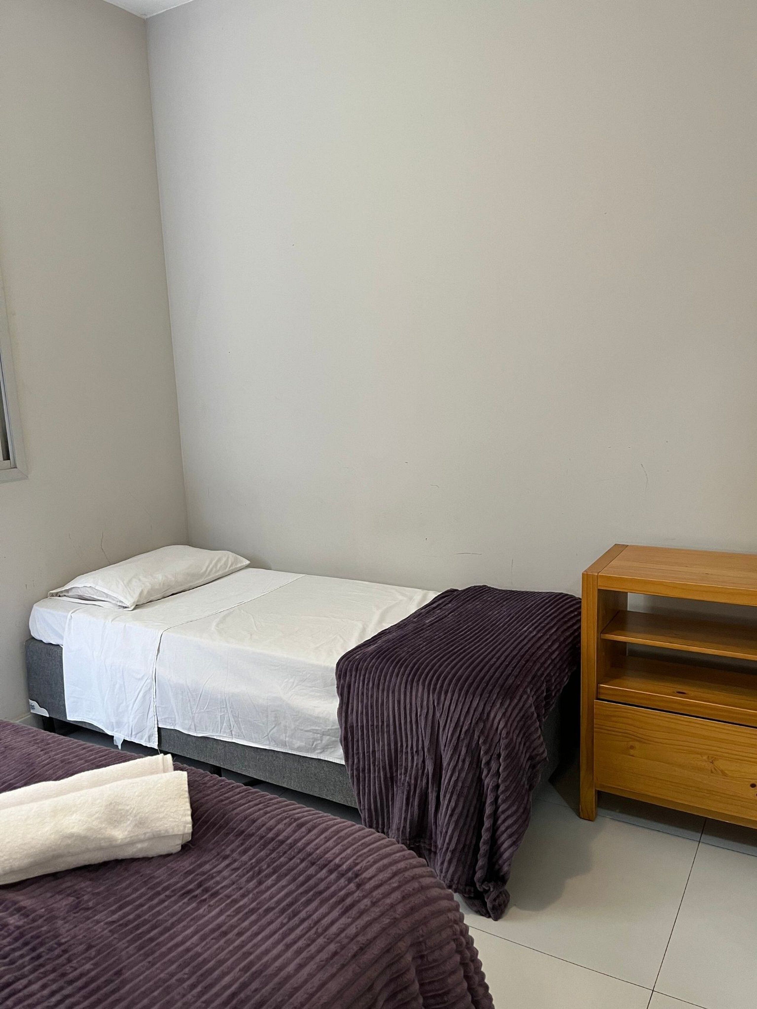 A small, clean room with a single bed, a wooden nightstand, and a window.
