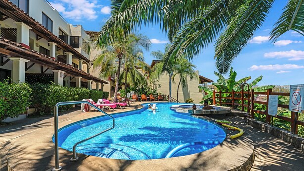 Une piscine entourée de chaises longues et de palmiers, le tout bordé d’un immeuble doté de balcons.