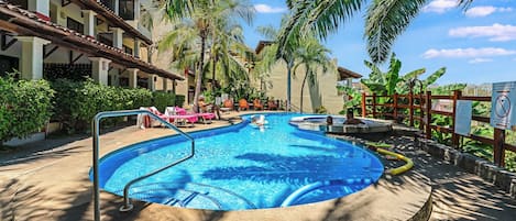 Une piscine entourée de chaises longues et de palmiers, le tout bordé d’un immeuble doté de balcons.