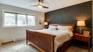 Une chambre à coucher comprenant un lit, une table de chevet, une lampe fixée au mur, un ventilateur de plafond et une fenêtre donnant sur des arbres.