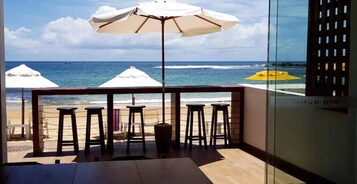 Un balcón con vista a la playa, sombrillas blancas y taburetes de bar.