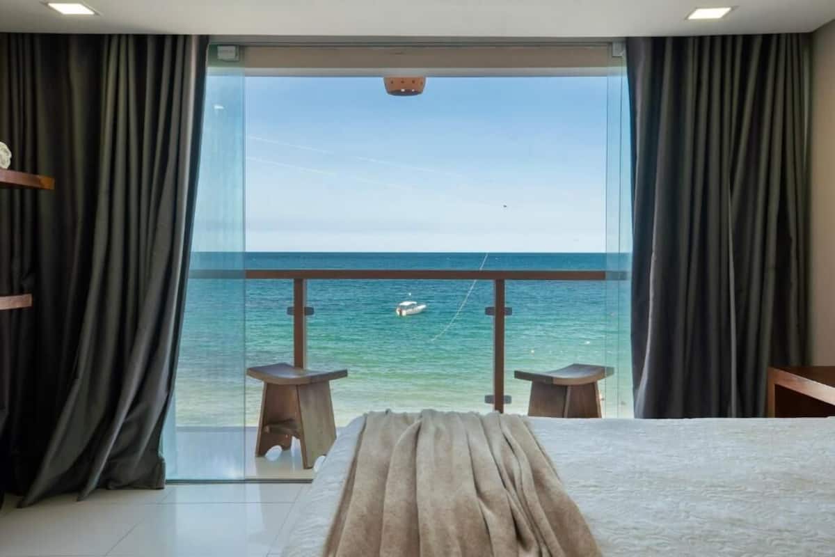 Un dormitorio con una cama grande, dos mesitas y vista al mar a través de un ventanal.