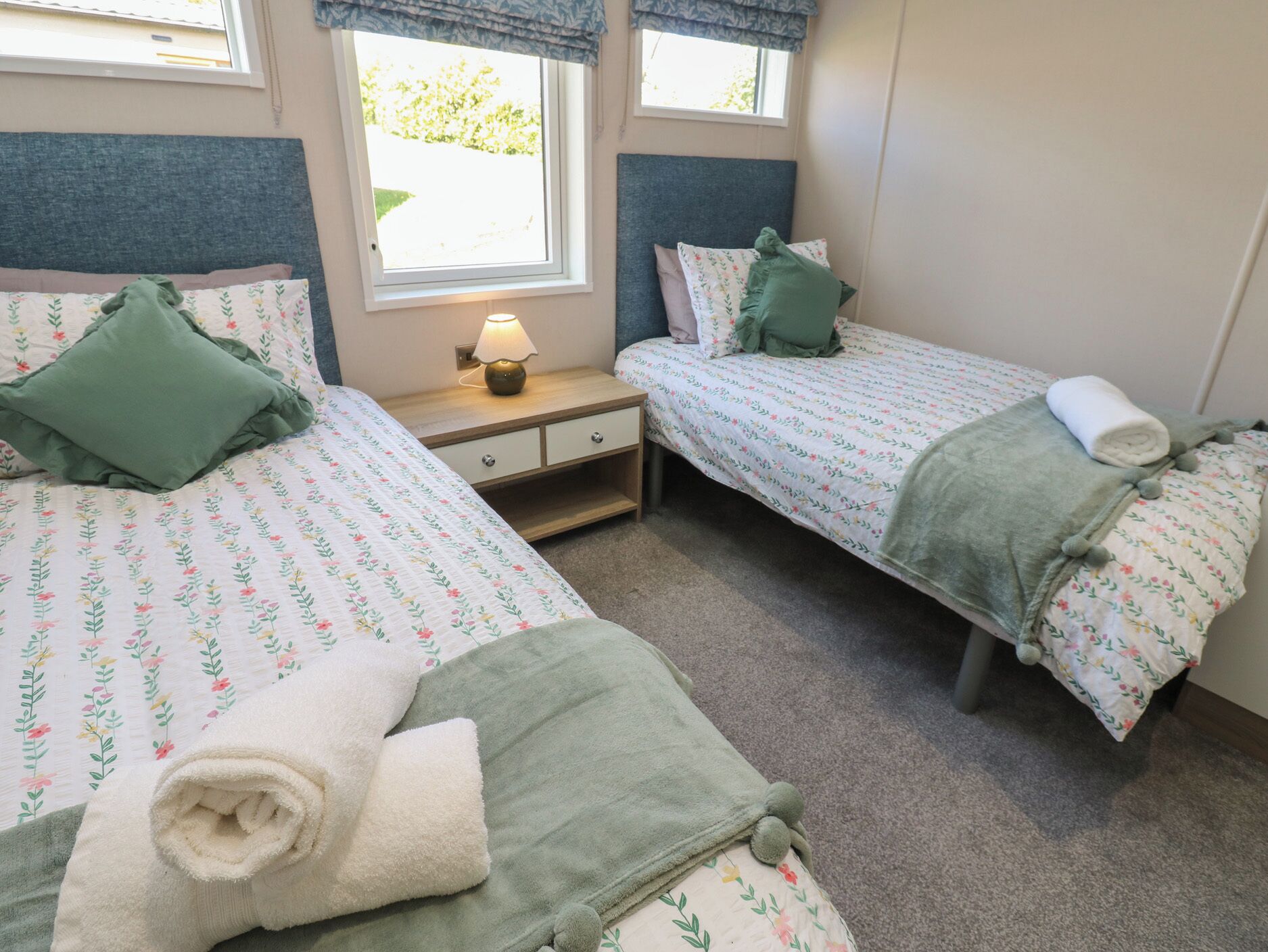 Habitación doble con dos camas, cada una con ropa de cama floral y almohadas verdes, una mesita de noche con lámpara, y una ventana con vistas a la vegetación.
