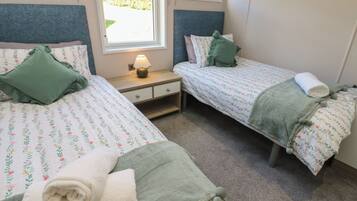 Habitación doble con dos camas, cada una con ropa de cama floral y almohadas verdes, una mesita de noche con lámpara, y una ventana con vistas a la vegetación.