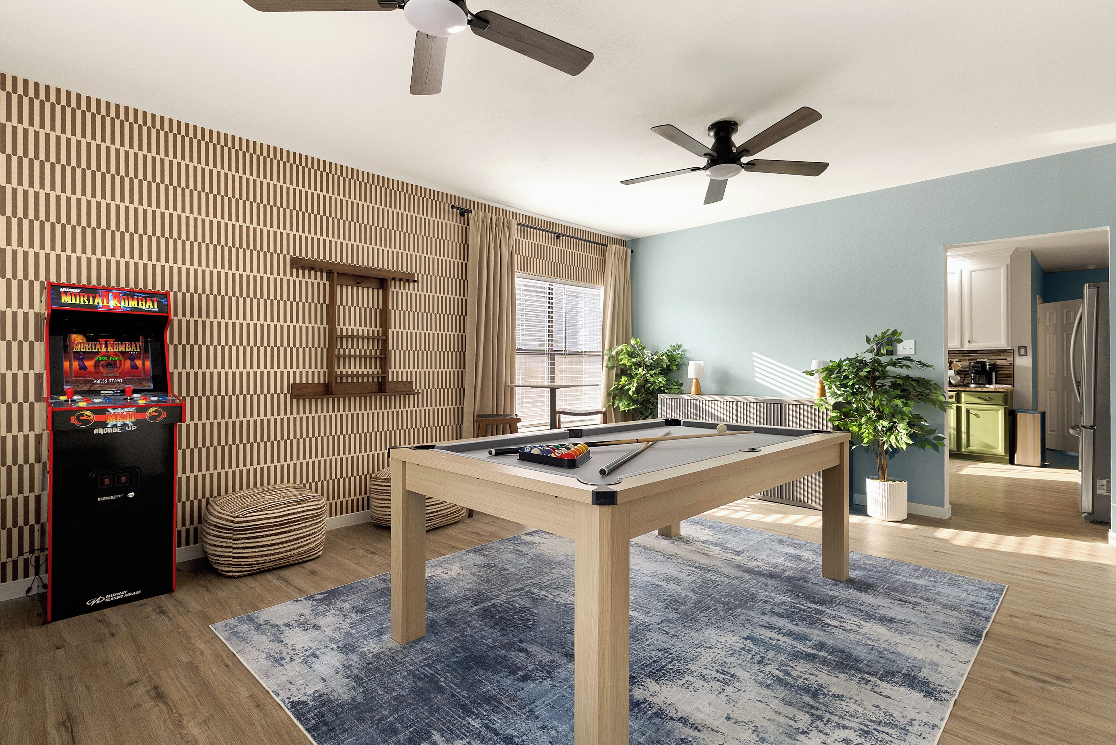 Ein Billardtisch mit blauem Teppich, einem Billardstöcke und einem Spielball. Es gibt einen Deckenventilator, ein an der Wand befestigtes Spielgerät und einen Blumentopf.