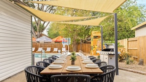 An outdoor dining area with a long table set for a meal, surrounded by chairs, under a shaded canopy. - The Saddle Club • Pool • Cinema • Vibes • Arcade (San Antonio)