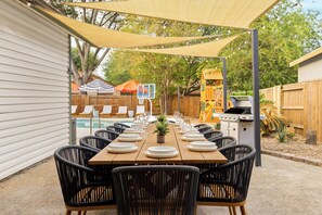 Un área de comedor al aire libre con una larga mesa dispuesta para una comida, rodeada de sillas, bajo un toldo sombreado.