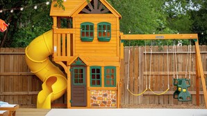 A wooden playhouse with a slide, windows, and a swing set. - The Saddle Club • Pool • Cinema • Vibes • Arcade (San Antonio)
