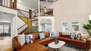 A spacious living room with a large sectional sofa, a wooden staircase, and a view of the outdoors. - The Saddle Club • Pool • Cinema • Vibes • Arcade (San Antonio)