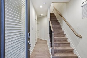 A staircase with wooden steps and a metal handrail, leading to an interior space with white walls and a door with a security lock. - Gated Modern Home in Independence Heights! (Houston)