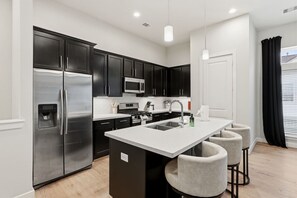 A modern kitchen with stainless steel appliances, a central island with bar stools, and a view of the outside through a window with curtains. - Gated Modern Home in Independence Heights! (Houston)