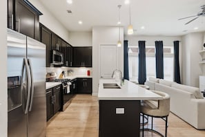 A modern kitchen with dark cabinets, stainless steel appliances, a central island, and a dining area with a white sofa. - Gated Modern Home in Independence Heights! (Houston)