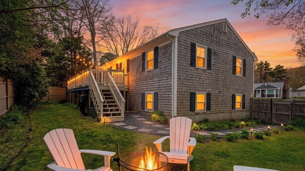 A two-story house with a fire pit in the backyard, surrounded by white Adirondack chairs. - Walk to 4 Beaches · Pool Table · Xbox · Fire Pit (Barnstable)