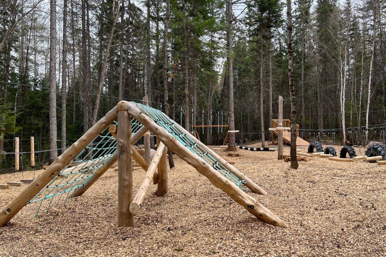 Ein Spielplatz mit einer hölzernen Kletterkonstruktion und einer Rutsche, umgeben von einem bewaldeten Gebiet.