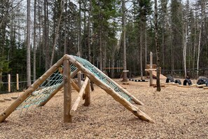 A playground with a wooden climbing structure and a slide, surrounded by a forested area. - La Sieste | Chalet avec SPA, plage et nature (Sainte-Lucie-des-Laurentides)
