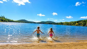 Two children playing in shallow water near a sandy shore with a clear sky and a forested hill in the background.
