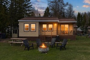 Een klein houten huisje met een terras, buitenmeubels en een vuurkuil.