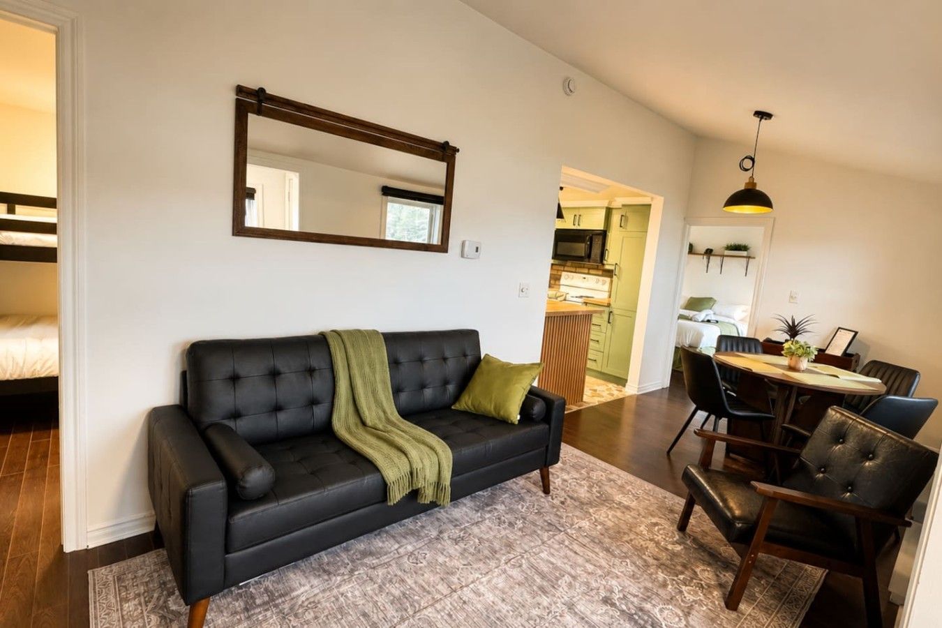 A modern living room with a black leather sofa, a dining area with a wooden table and chairs, and a kitchen visible through an open doorway.
