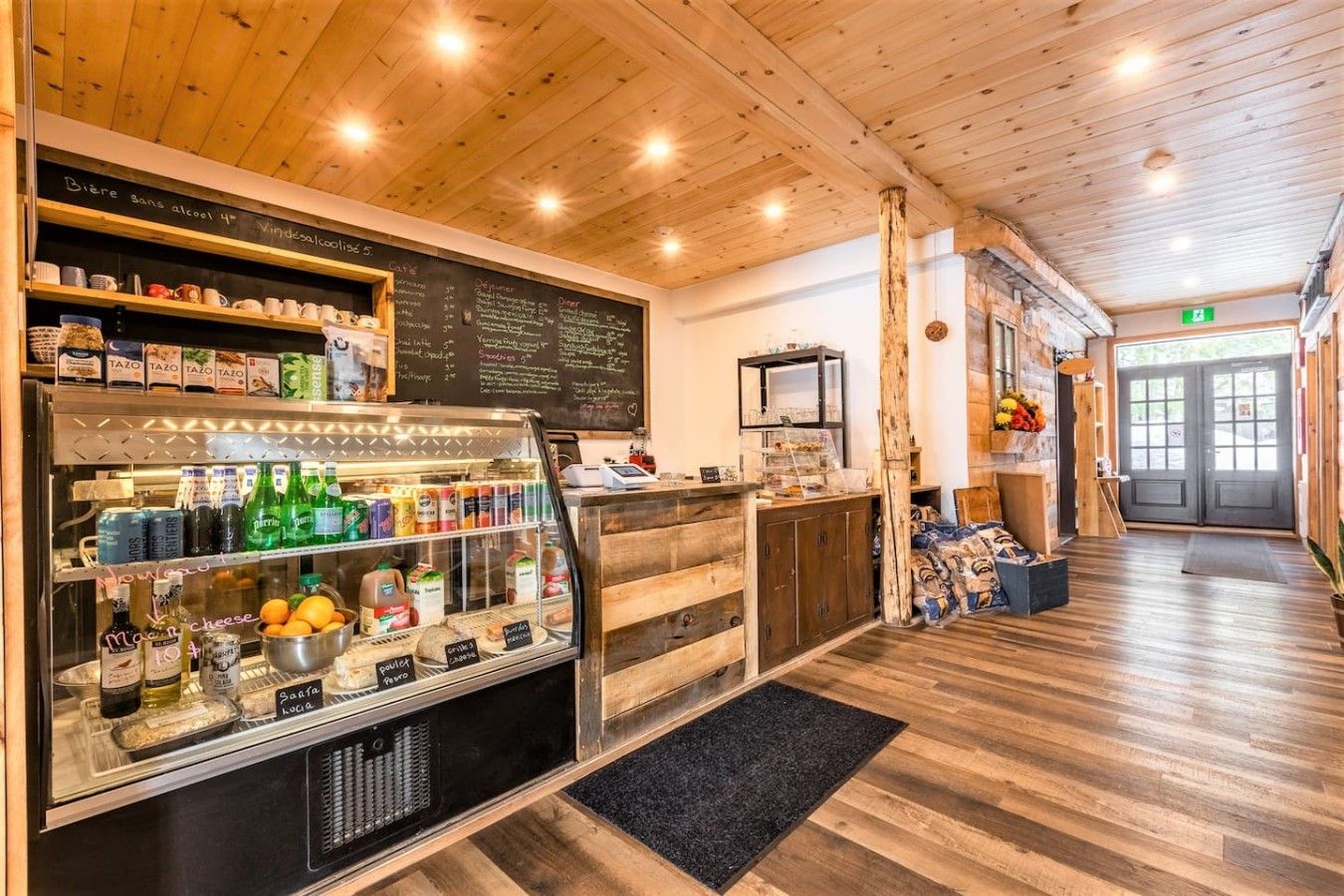 Interior de una cafetería con techo de madera, un menú en una pizarra y una barra con diversas bebidas y aperitivos.