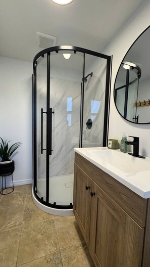 Un baño moderno con ducha envuelta en cristal, tocador de madera y espejo redondo.