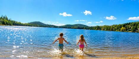 Two children playing in shallow water near a sandy shore with a clear sky and a forested hill in the background.