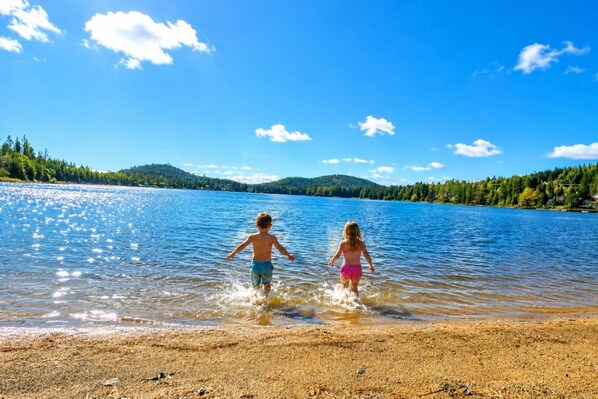 Dos niños jugando en aguas poco profundas cerca de una orilla arenosa, bajo un cielo despejado y con una colina boscosa al fondo.